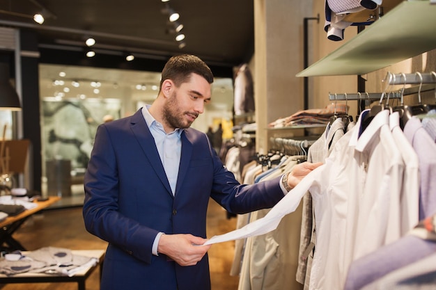 Venda, compras, moda, estilo e conceito de pessoas - jovem feliz de terno escolhendo camisa no shopping ou loja de roupas