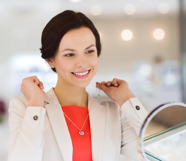 Venda, consumismo, compras e conceito de pessoas - mulher feliz escolhendo e experimentando pingente na joalheria