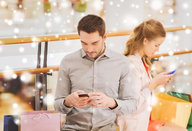 venda, consumismo, tecnologia e conceito de pessoas - jovem casal com sacolas de compras e smartphones mensagem de texto no shopping com efeito de neve