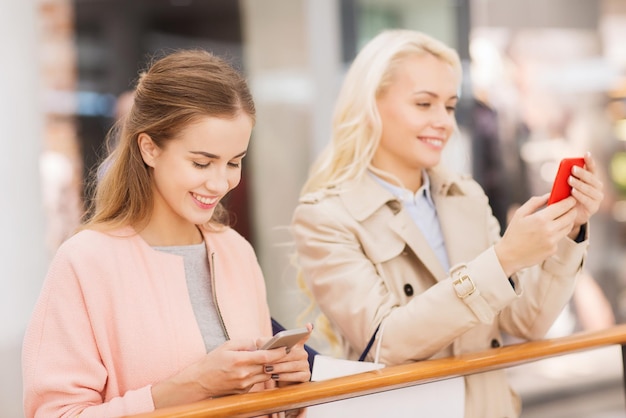 Venda, consumismo, tecnologia e conceito de pessoas - mulheres jovens felizes com smartphones e sacolas de compras no shopping