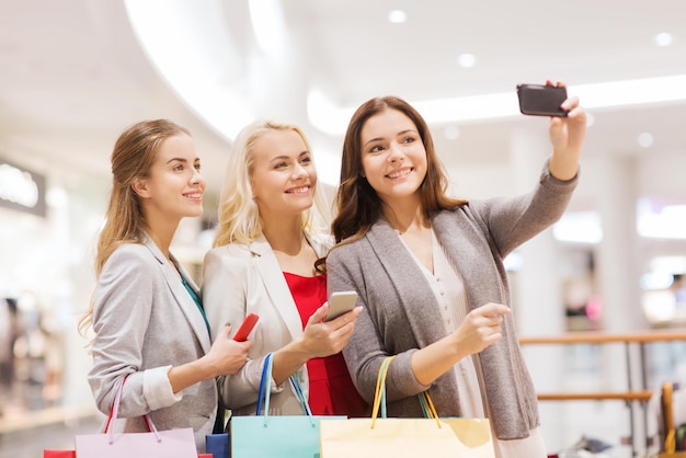 Venda, consumismo, tecnologia e conceito de pessoas - mulheres jovens felizes com smartphones e sacolas de compras tomando selfie no shopping