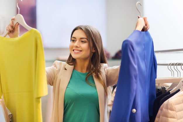 venda, roupas, compras, moda e conceito de pessoas - jovem feliz escolhendo entre camisa e jaqueta na loja de roupas