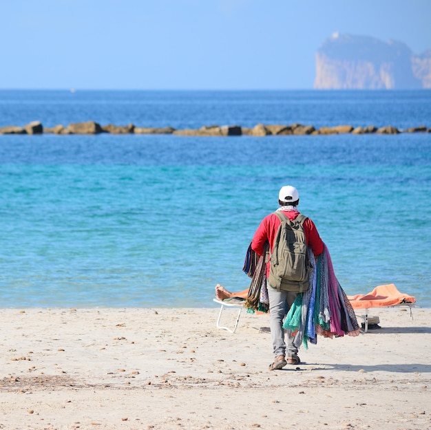 Vendedor andando na praia na costa de Alghero Capo Caccia no fundo em um dia ensolarado
