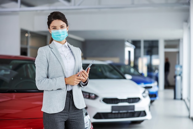 Vendedor de autos femenino en traje con mascarilla de pie en el salón del automóvil y sosteniendo la tableta durante el brote del virus corona.