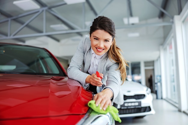 Vendedor de coches femenino ordenado sonriente frotando el coche con detergente y paño. Todo debe estar brillante y limpio.