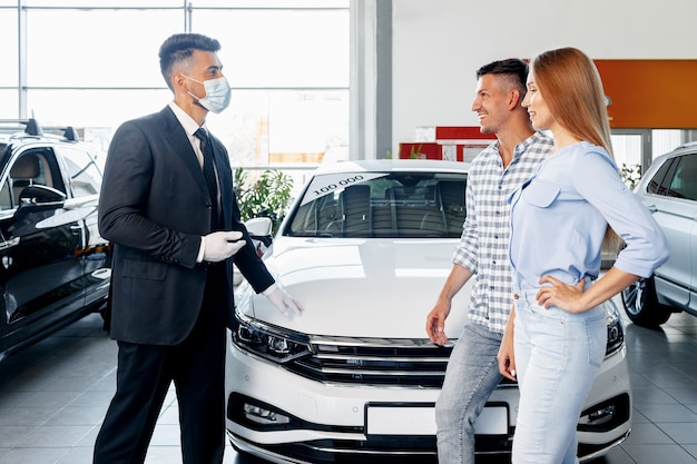 Vendedor de coches de hombre en mascarilla hablando con un cliente en la sala de exposición de coches