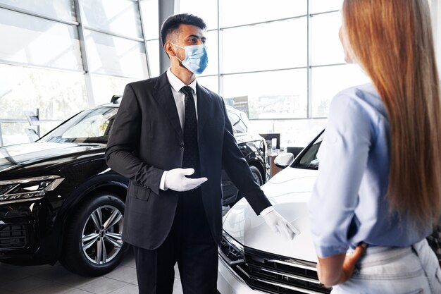 Vendedor de coches de hombre en mascarilla hablando con un cliente en la sala de exposición