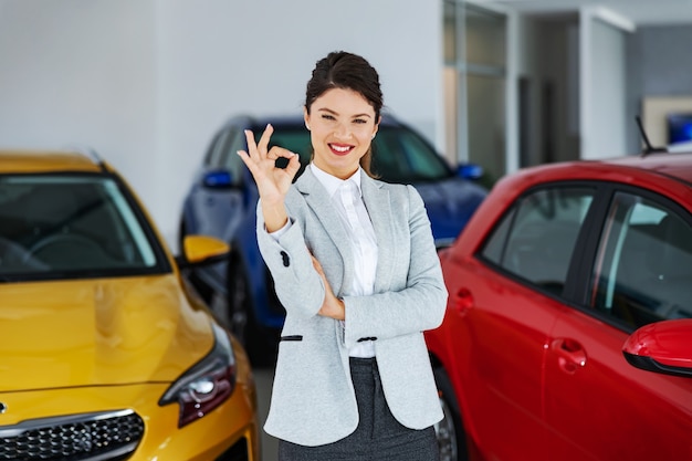 Vendedor de coches mujer sonriente de pie en el salón del coche y mostrando signo bien.