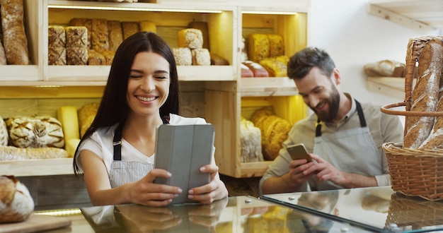 Vendedor de mulher bonita caucasiana, rolagem e gravação no computador tablet em pé no balcão da loja de padaria, homem falando ao telefone atrás dela. colegas de trabalho conversando e rindo. interior