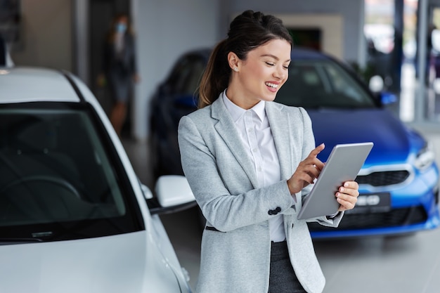 Vendedor feminino sorridente de terno usando tablet para procurar qual carro é vendido em pé no salão de beleza.