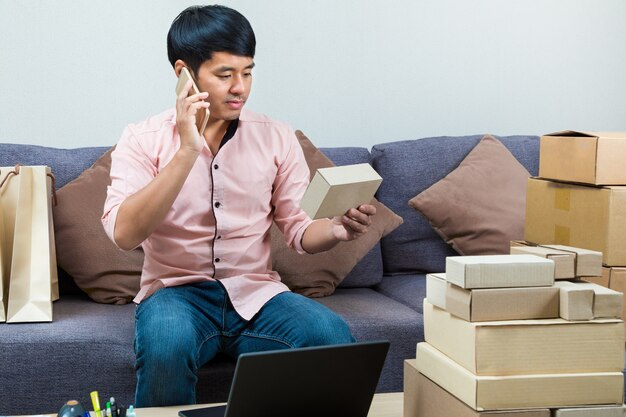 Vendedor en línea asiático masculino que habla en el teléfono mientras que sostiene una caja de su producto