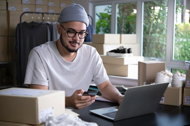 Vendedor en línea de hombre asiático que confirma pedidos del cliente por teléfono Propietario de negocio masculino de comercio electrónico mirando el teléfono en el almacén de la tienda