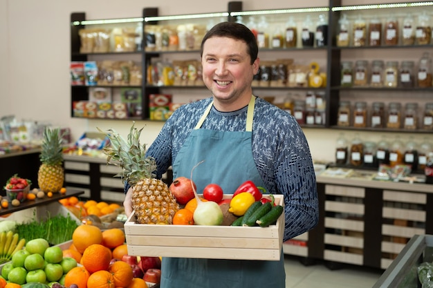 Vendedor masculino sorridente segura uma caixa de madeira com legumes e frutas na loja.