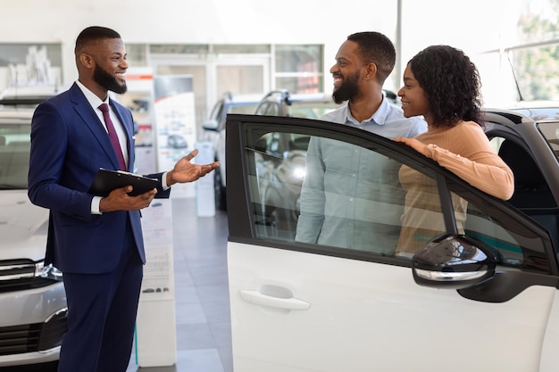 Vendedor no centro de concessionárias de automóveis ajudando os cônjuges negros a escolher o novo veículo da família, explicando as características do automóvel para o jovem casal afro-americano em pé no showroom de automóveis