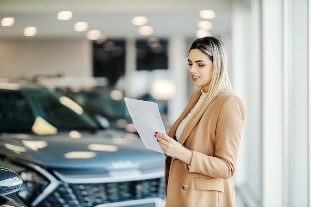 Una vendedora de autos feliz está parada en la sala de exhibición con papeleo en sus manos y mirándolo