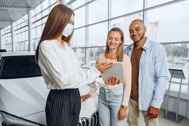 Vendedora de coches con máscara médica muestra a los compradores algo en tableta digital