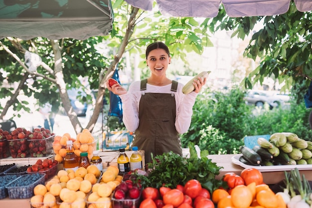 Foto vendedora joven que sostiene el calabacín y el tomate en manos