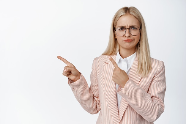 Vendedora preocupada con gafas y traje apuntando con el dedo hacia la izquierda, frunciendo el ceño y mirando dudosa, quejándose de algo en blanco.