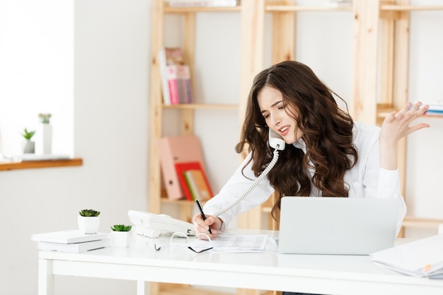 Vendedora séria e bem-vestida falando ao telefone no escritório atrás de sua mesa e copiando o espaço do laptop