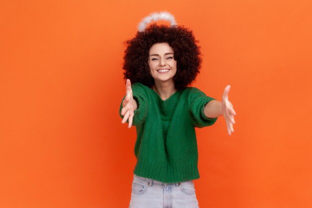 Foto venha para os meus braços. mulher angelical sorridente com penteado afro no suéter verde estilo casual e nimb sobre a cabeça esticando as mãos, quer abraçá-lo. tiro de estúdio interior isolado em fundo laranja.