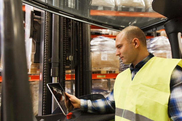 venta al por mayor, logística, carga, envío y concepto de personas: hombre o cargador con computadora de tableta y carretilla elevadora o cargador cargando cajas en el almacén