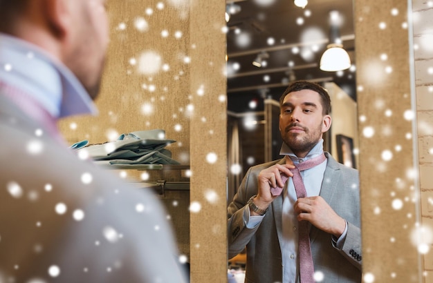 venta, compras, moda, estilo y conceptos de personas - joven feliz eligiendo y atando corbata y mirando al espejo en el centro comercial o tienda de ropa sobre la nieve