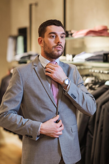 venta, compras, moda, estilo de negocios y concepto de personas - joven elegante eligiendo y probándose traje y corbata en un centro comercial o tienda de ropa