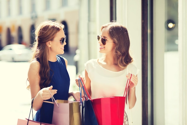 venta, consumismo y concepto de personas - mujeres jóvenes felices con bolsas de compras hablando en la ventana de la tienda en la ciudad