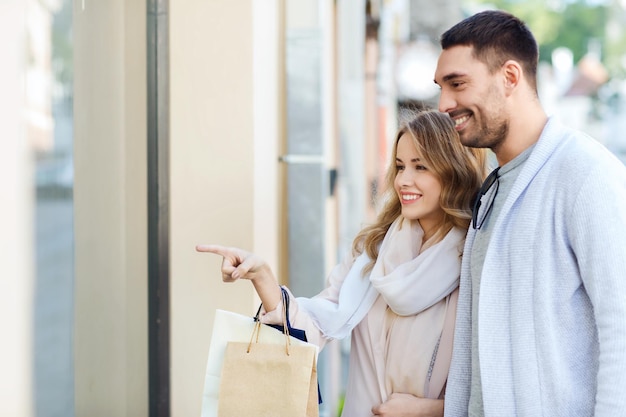 venta, consumismo y concepto de personas - pareja feliz con bolsas de compras apuntando con el dedo a la ventana de la tienda en la calle de la ciudad