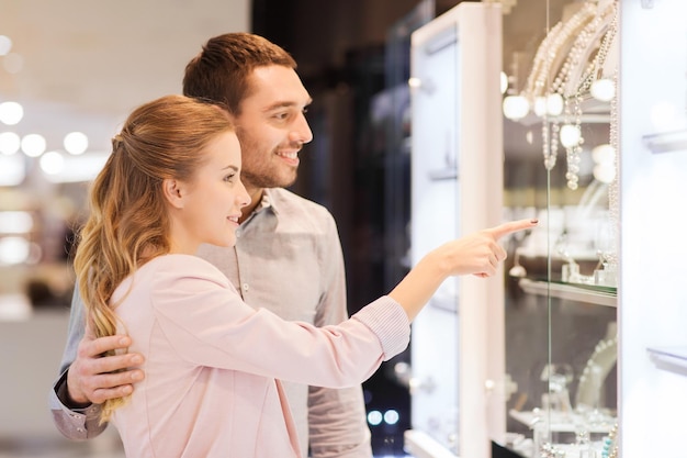 venta, consumismo y concepto de personas - pareja feliz señalando con el dedo a la ventana de compras en la joyería en el centro comercial