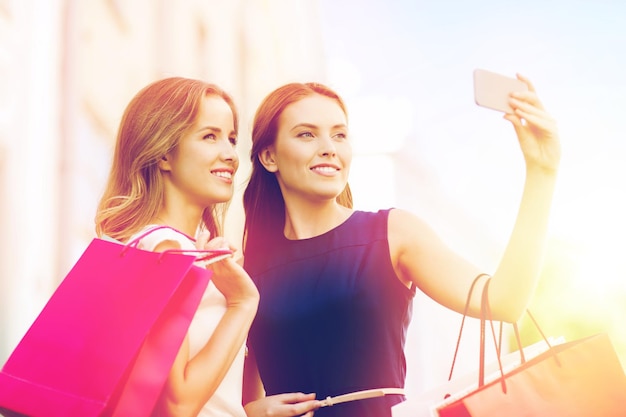 Venta, consumismo, tecnología y concepto de personas: mujeres jóvenes felices con bolsas de compras y teléfonos inteligentes tomando selfie en la calle de la ciudad