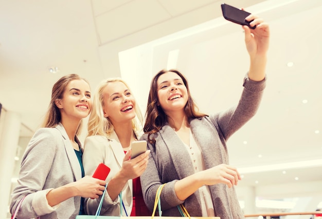 venta, consumismo, tecnología y concepto de personas - mujeres jóvenes felices con smartphones y bolsas de compras tomando selfie en el centro comercial