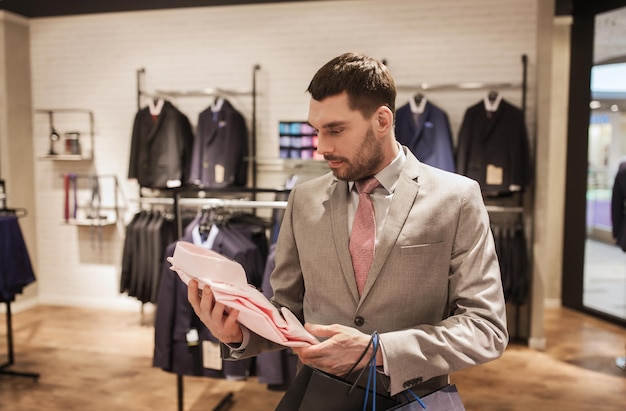 venta, moda, estilo y concepto de la gente - elegante joven o hombre de negocios en traje con bolsas de compras eligiendo camisa en el centro comercial o tienda de ropa