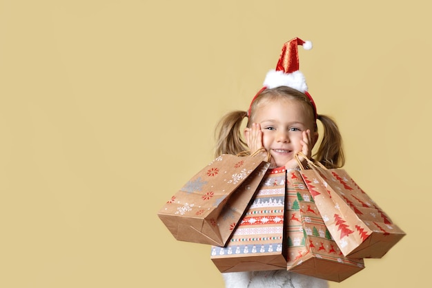 Venta de Navidad. Sorprendido niño feliz niña con sombrero de Santa sosteniendo bolsas de papel artesanales de regalo de compras presentes estudio de fondo amarillo aislado descuento en línea estacional. Simulacros, espacio de copia, banner. Viernes negro