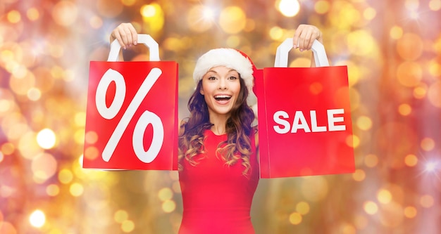 venta, navidad, vacaciones y concepto de la gente - mujer joven sonriente feliz en sombrero de santa sosteniendo bolsas de compras rojas con signo de porcentaje sobre fondo de luces