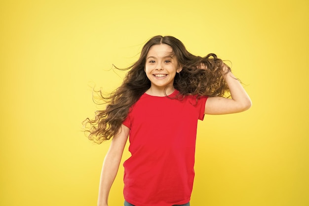 Ventos fortes e persistentes podem criar emaranhados e emaranhados em cabelos longos ondulados e cacheados Coisas que você faz para danificar seu cabelo Garota adorável criança cabelo longo ondulado fundo amarelo O vento também pode danificar o cabelo