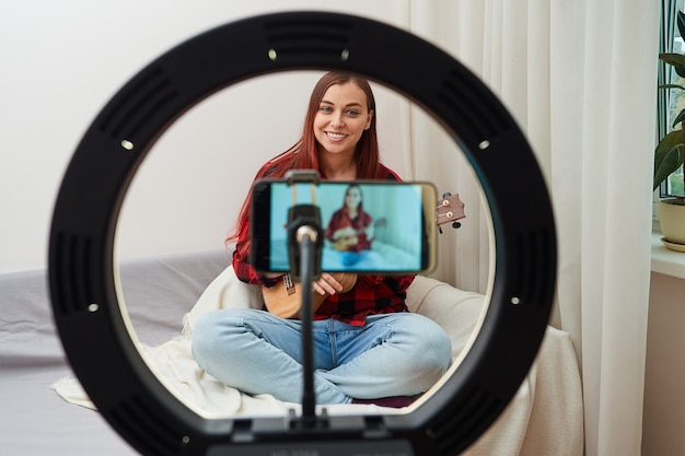 Foto ver através de uma lâmpada de anel músico blogueiro grava uma transmissão online em um telefone celular em que uma mulher toca ukulele para seus seguidores