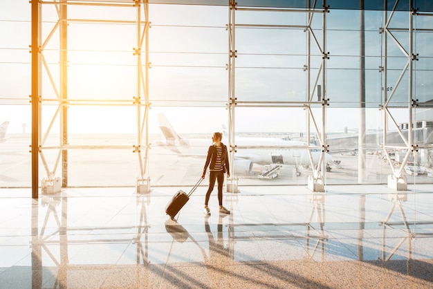 Ver os na janela do aeroporto com mulher andando com mala no salão de embarque durante o pôr do sol. Visão de grande angular com espaço de cópia