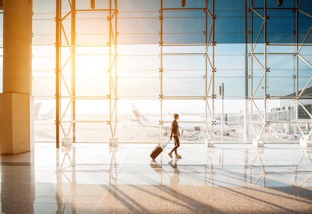 Ver os na janela do aeroporto com mulher andando com mala no salão de embarque durante o pôr do sol. Visão de grande angular com espaço de cópia