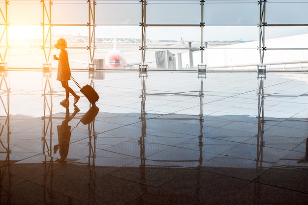 Ver os na janela do aeroporto com mulher andando com mala no salão de embarque durante o pôr do sol. Visão de grande angular com espaço de cópia