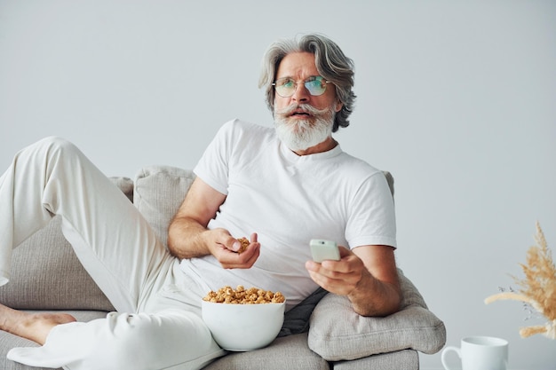 Ver programa de televisión y comer palomitas de maíz Hombre moderno con estilo senior con cabello gris y barba en el interior