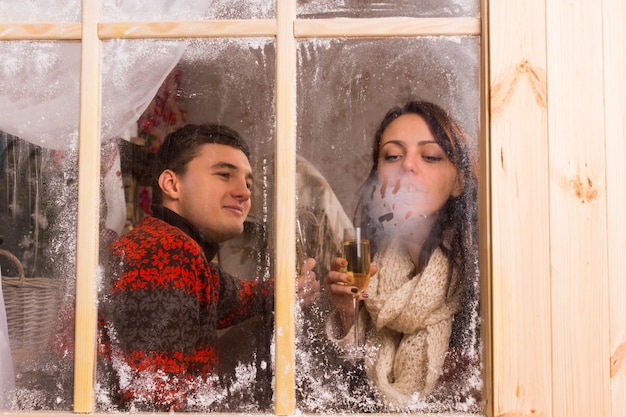 Ver a través de la fría ventana esmerilada de una joven pareja celebrando en una cabaña de invierno disfrutando de una bebida y soplando sobre el vidrio para condensar su aliento en el frío