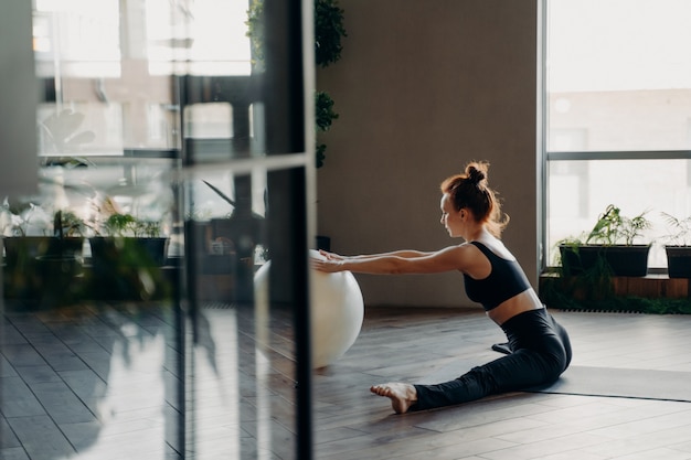 Ver a través de la puerta de vidrio abierta de una mujer delgada pelirroja flexible en ropa activa sentada en un cordel en la estera con las manos en la gran fitball mientras realiza ejercicios de estiramiento en el gimnasio o estudio de fitness