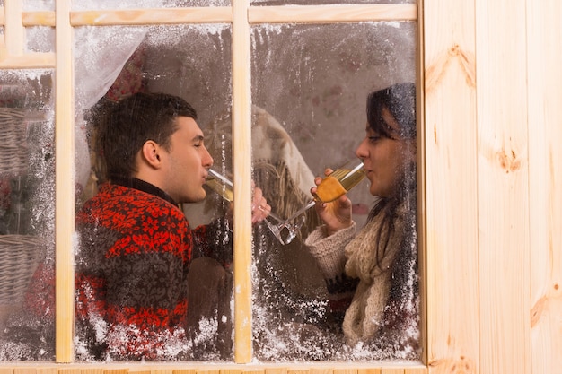 Ver a través de una ventana de madera rústica helada Pareja joven celebrando en Navidad bebiendo copas de champán mientras están uno frente al otro