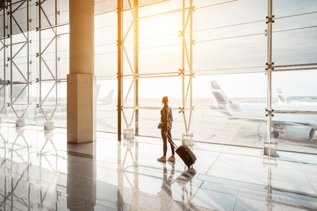 Ver en la ventana del aeropuerto con una mujer caminando con maleta en la sala de salidas durante la puesta de sol. Vista de gran angular con espacio de copia