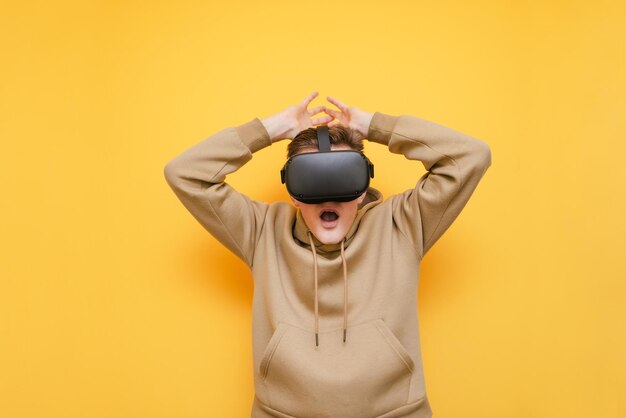 Verängstigter junger Mann, der in einer VR-Brille auf gelbem Hintergrund steht und mit schockiertem Gesicht in Freizeitkleidung in die Kamera schaut Porträt eines ausdrucksstarken Mannes in einem VR-Helm, der vom Realismus überrascht ist