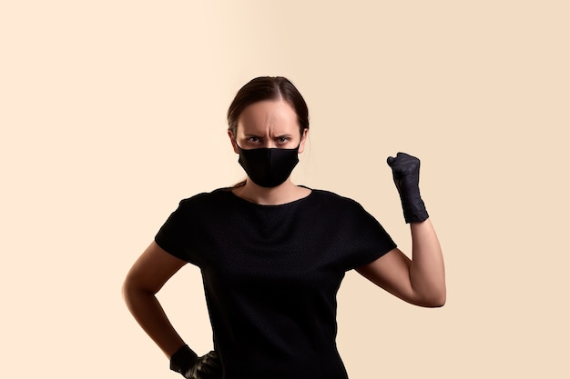 Verärgerte Frau im schwarzen Kleid Gesichtsmaske und Latexhandschuhe, die Faust und über beige Wand zeigen