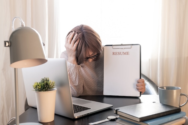 Verärgerte Frau schloss Ihr Gesicht der Hände in der Nähe ihres Arbeitsplatzes mit Laptop und Lebenslauf Anwendung.