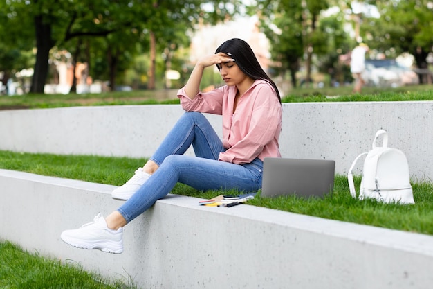 Verärgerte hispanische Dame fühlt sich müde, während sie nach dem Unterricht mit Laptop im Freien lernt und im Park sitzt