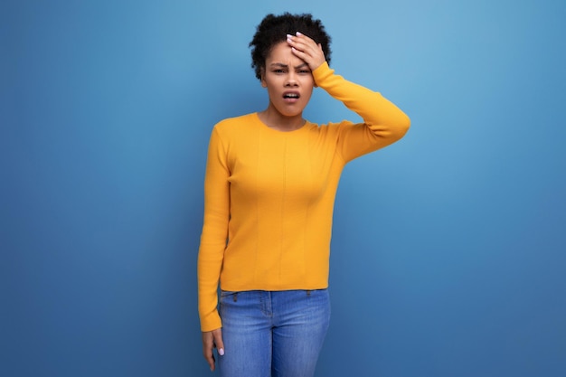 Verärgerte hübsche junge hispanische Frau mit schwarzem lockigem Haar in einem gelben Pullover auf blauem Hintergrund